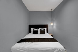 A small, narrow hotel room with a single bed, a nightstand, and a wall-mounted light fixture.