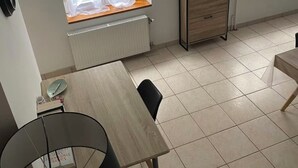 Huoneessa on ruokapöytä, kaksi ikkunaa, joissa on ohuet verhot, patteri ja pieni puinen kaappi.