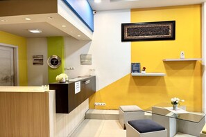 A reception area with a wooden reception desk, a green accent wall, a yellow accent wall, a framed calligraphy piece, a small shelf with a vase, and a glass table with a vase.