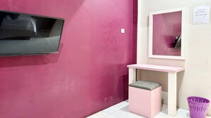 Una habitación con una pared rosa, un televisor de pantalla plana montado en la pared, un escritorio rosa pequeño con una silla y una cesta morada en el suelo.