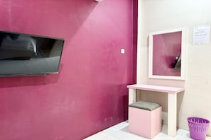 Una habitación con una pared rosa, un televisor de pantalla plana montado en la pared, un escritorio rosa pequeño con una silla y una cesta morada en el suelo.