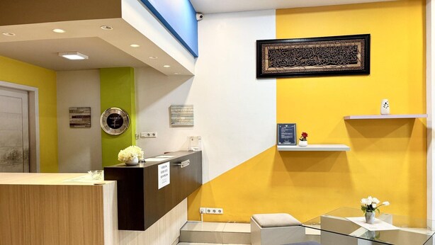 Zona de recepción con mostrador de madera, pared de acento verde, pared de acento amarilla, una obra de caligrafía enmarcada, un estante con un jarrón y una mesa de cristal con un jarrón.