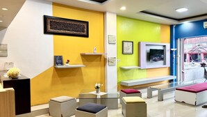 Un área de recepción con diseño moderno, que incluye un mostrador de recepción, una zona de estar con otomanas, un televisor montado y obras de arte en las paredes.