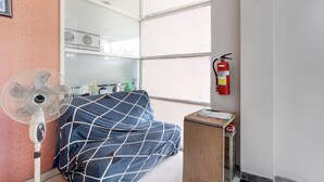 A room with a bed, a standing fan, a fire extinguisher, and a window with blinds.