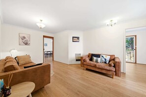 Een ruime woonkamer met een bruine bank, een houten salontafel, en een eethoek met een tafel en stoelen.