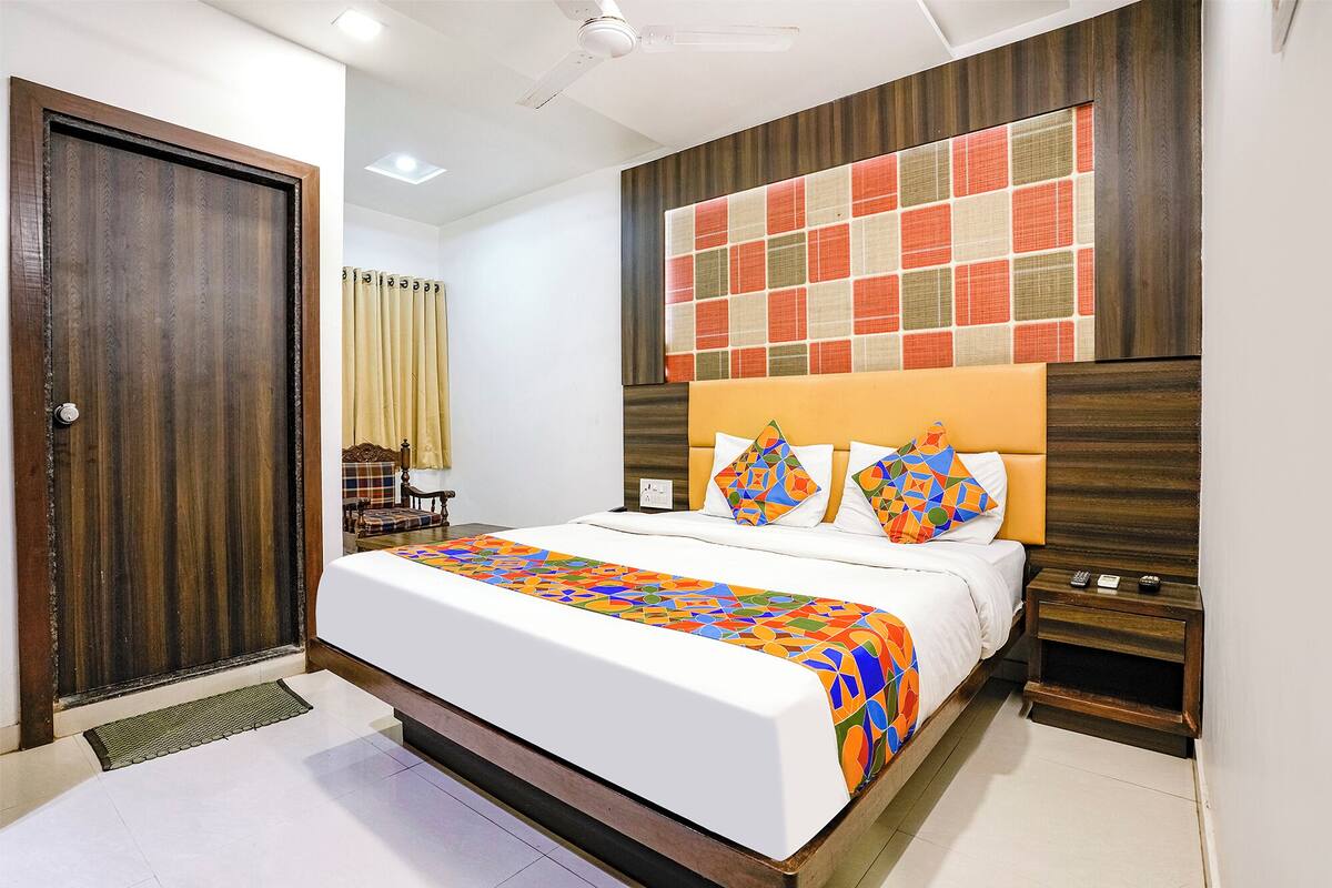 A hotel room with a bed, a wooden headboard, a nightstand, a chair, a window with curtains, and a door.