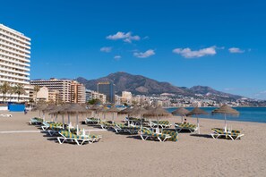 Een strand met rijen ligbedden en parasols, een hotel op de achtergrond en bergen in de verte.