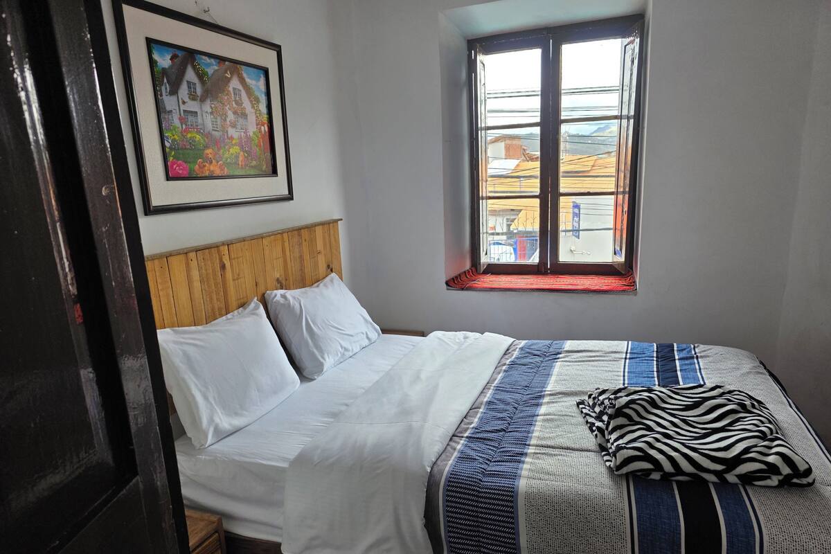 Un dormitorio con cabecera de madera, una cama individual con ropa de cama blanca, un edredón a rayas y una ventana con vistas a edificios.