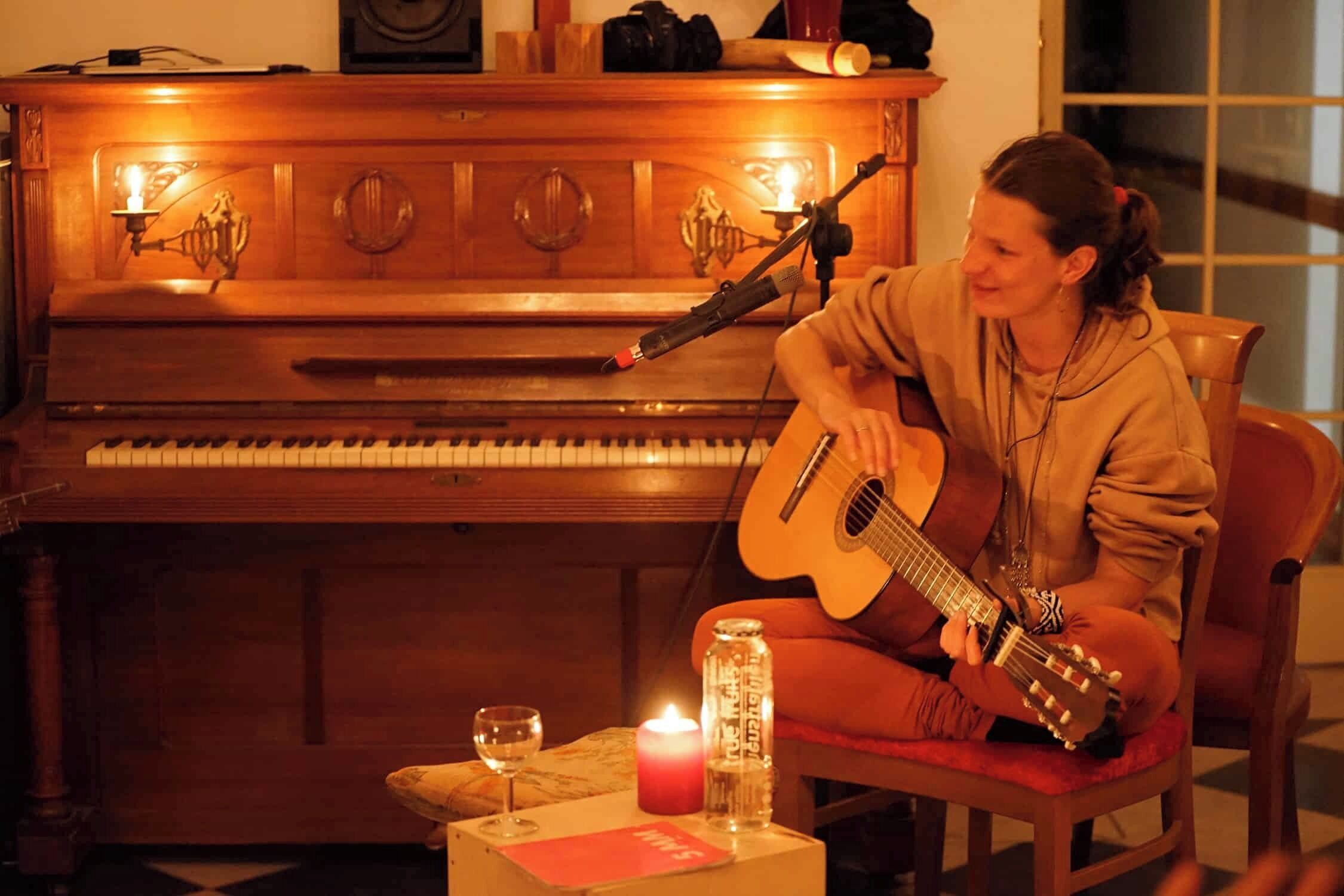 A person playing a guitar near a piano in a warmly lit room with a candle and a glass on a small table.