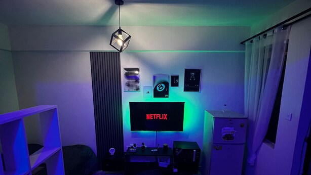 Una habitación pequeña y con poca luz, con un televisor mostrando 'NETFLIX', un escritorio con una computadora, una silla y una estantería con diversos objetos.