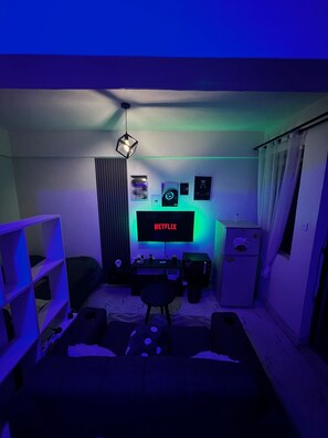 A small, dimly lit room with a TV displaying 'NETFLIX,' a desk with a computer, a chair, and a shelf with various items. - Top-Floor cozy  Studio with Stunning Views lower kabete (Nairobi)