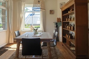 A dining room with a wooden table, chairs, a hanging light fixture, a window with curtains, and a wooden cabinet with kitchen items.