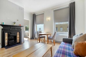 Un salon avec une cheminée, une table ronde entourée de chaises, un canapé et de grandes fenêtres avec des rideaux.