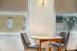Une pièce avec une table ronde en bois et deux chaises grises à revêtement rembourré, de grandes fenêtres donnant sur un bâtiment et une plante à l’extérieur.