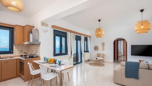 A modern kitchen with a dining area, a living room with a sofa and TV, and large windows with curtains. - Sacada Seascape Villa (Nisiros)