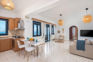 A modern kitchen with a dining area, a living room with a sofa and TV, and large windows with curtains.