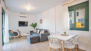 Een moderne woonkamer met een grijze bank, een witte eettafel en stoelen, een klein bijzettafeltje, een potplant en een wandtelevisie.