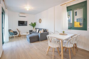 Ein modernes Wohnzimmer mit grauem Sofa, weißem Esstisch und Stühlen, einem kleinen Beistelltisch, einer Topfpflanze und einem Wand-Fernseher.