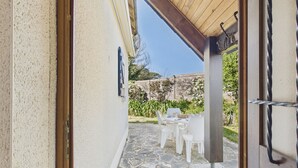 A stone patio with white chairs and a table, a wooden pergola, and a view of a garden.
