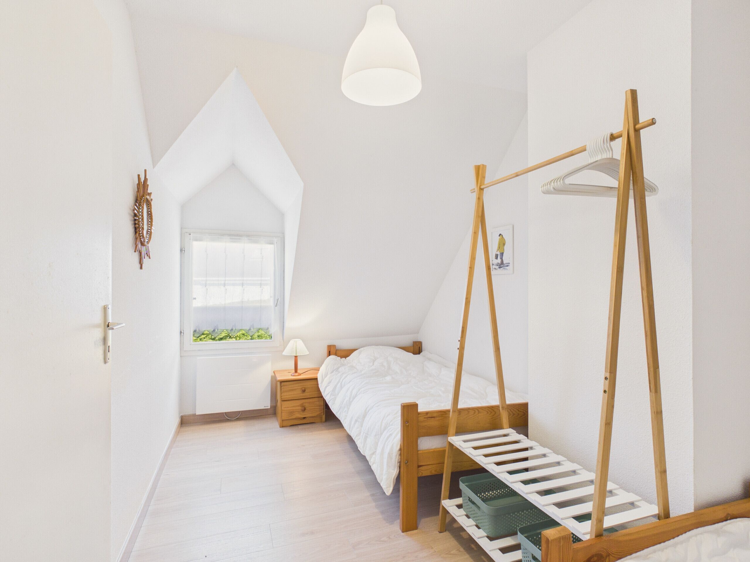A bedroom with a sloped ceiling, a bed with a wooden frame, a bedside table, a hanging light fixture, and a window with a view of greenery.