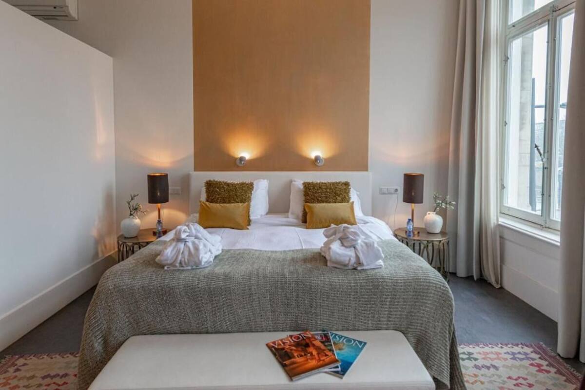 A hotel room with a bed, two bedside lamps, a window with curtains, and a bedside table with a lamp and a vase.