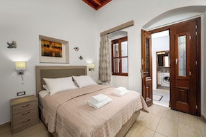 Un dormitorio con cama, mesitas de noche, un cuadro, una ventana con cortinas y una puerta abierta al baño.