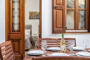 A dining table set with plates, glasses, and a bottle, with wooden chairs and a window with wooden shutters. - Villa Yannis in Lindos (Rhodes)