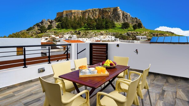 A terrace with a table set for breakfast, yellow chairs, and a view of a castle on a hill. - Villa Yannis in Lindos (Rhodes)