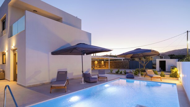 A modern outdoor pool area with lounge chairs and umbrellas, surrounded by a white building with large windows.