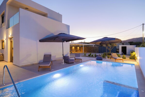 A modern outdoor pool area with lounge chairs and umbrellas, surrounded by a white building with large windows.