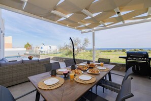 An outdoor dining area with a view of the sea, a grill, and a wicker sofa.
