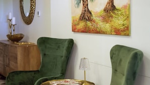 Een kamer met twee groene fauteuils, een zwart-wit vloerkleed, een schilderij van bomen, een spiegel en een tafeltje met een lamp.