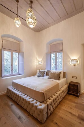 A bedroom with a large bed, two wall sconces, and a window with shutters. - Aylin Executive Suites - Sachnisi Suite (Rhodes)