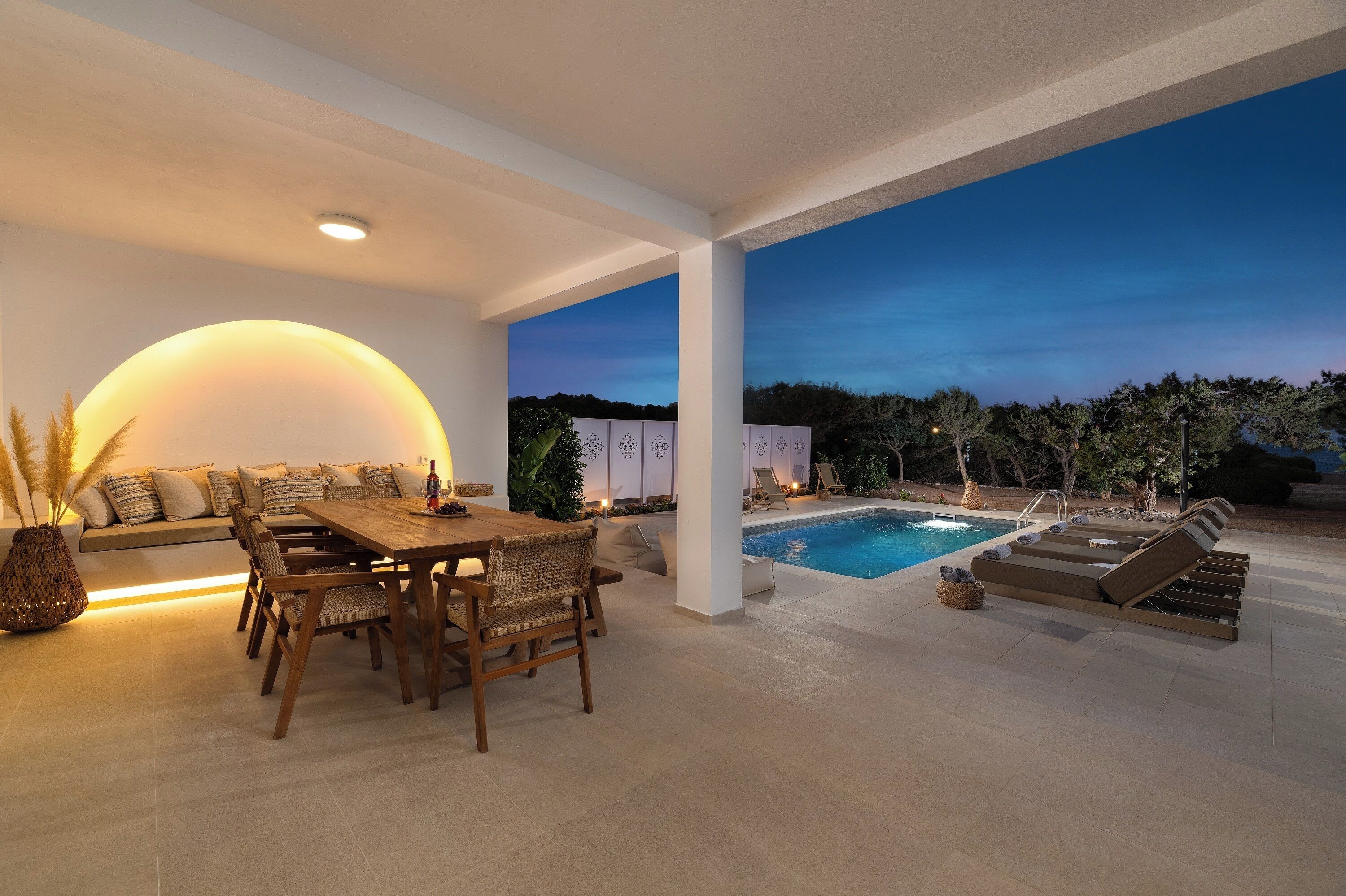 Uma área de estar moderna ao ar livre, com piscina, mesa de jantar com cadeiras e um espaço de descanso com sofá e almofadas.
