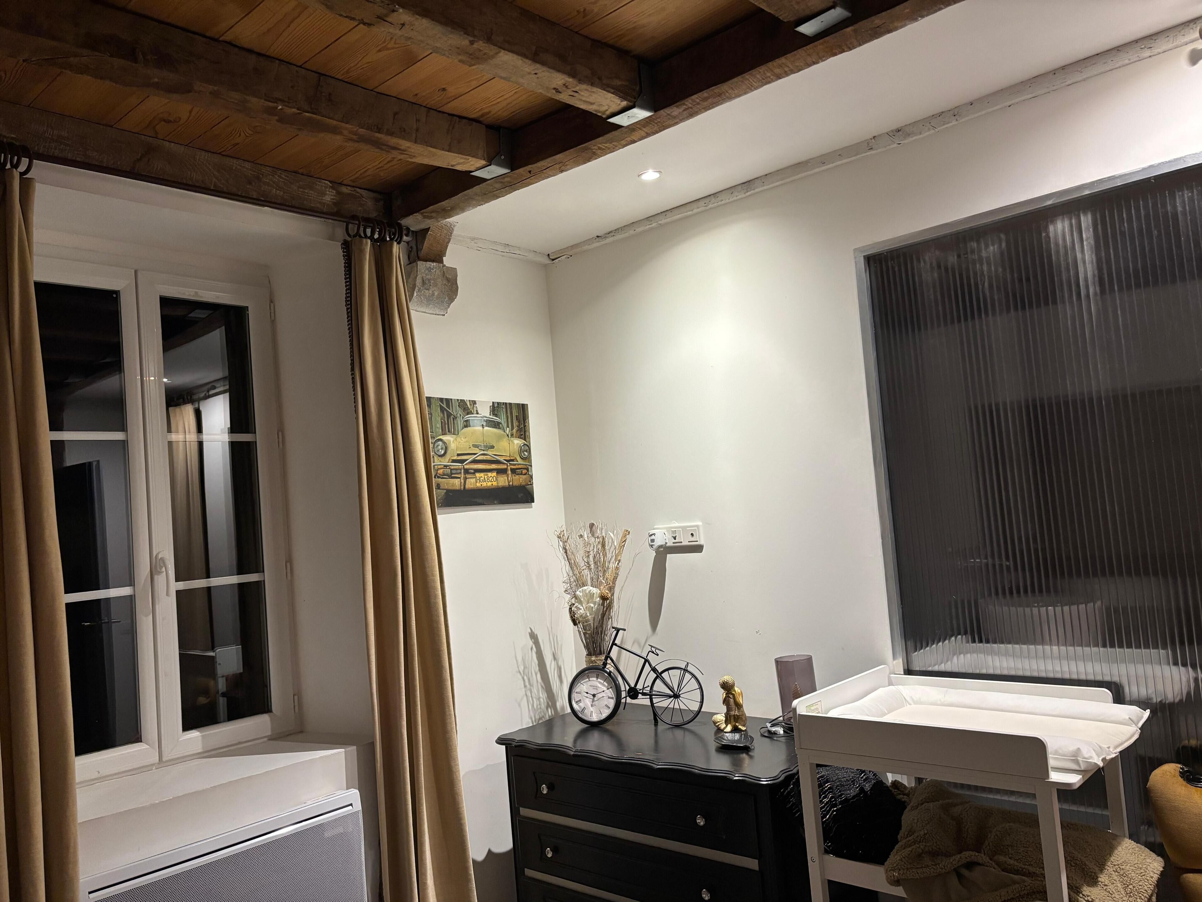 A bedroom with a window, a bedside table with a clock, a small bedside table with a teddy bear, a radiator, and a wall-mounted power outlet.