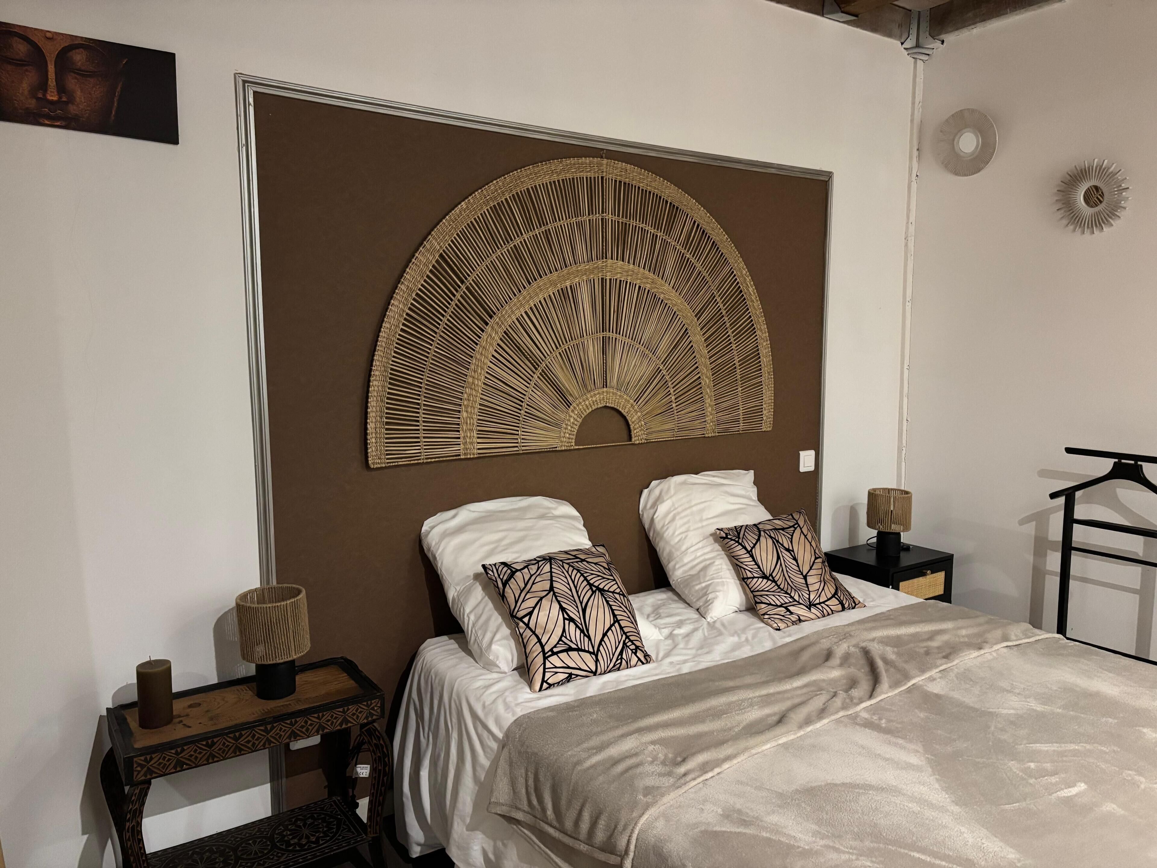 Un dormitorio con una cama grande, dos almohadas, una mesita de noche con lámpara y un cabecero decorativo fijado a la pared.
