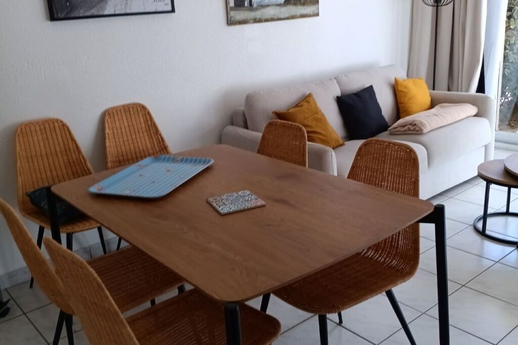 Een eetgedeelte met een houten tafel en rieten stoelen, een bank met kussens, en een raam met gordijnen.