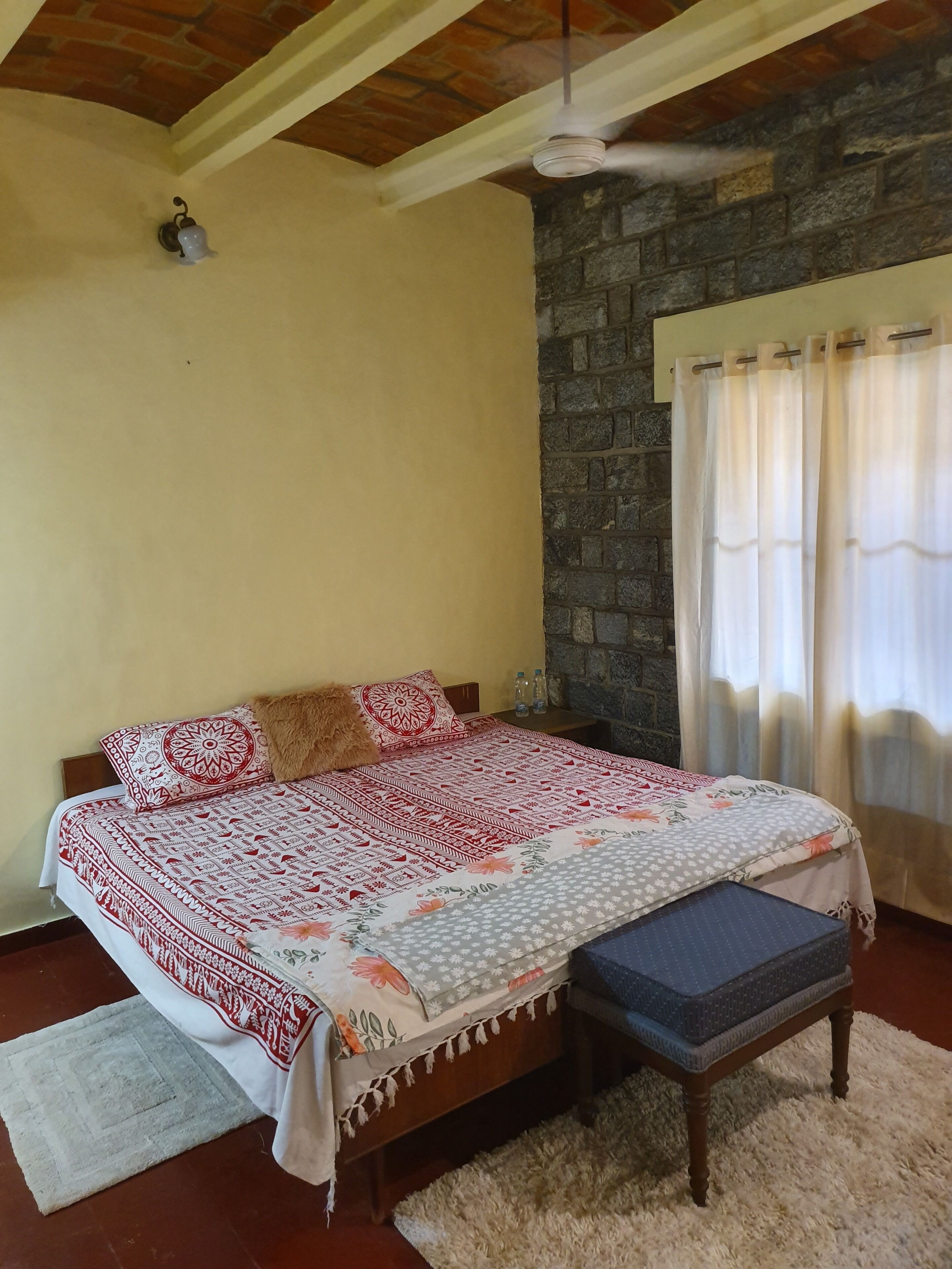 Quarto com cama, um banquinho, ventoinha no teto, parede de pedra e janela com cortinas.