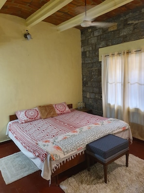 A bedroom with a bed, a bench, a ceiling fan, a stone wall, and a window with curtains.