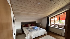 Un dormitori amb un llit, una tauleta de nit, una finestra amb un para-sol i bigues de sostre de fusta.
