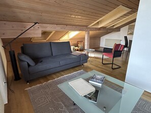 An attic room with a grey sofa, a glass coffee table with books, a standing lamp, and a radiator. - Apartment 57 sqm, balcony and separate bedroom (Grassau)