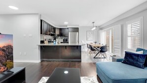A modern kitchen with dark cabinetry, a dining area with a table and chairs, and a blue sofa in a living room with a flat-screen TV. - Entire2Storey+Trampoline+65"+EnclosedYard (Kitchener)