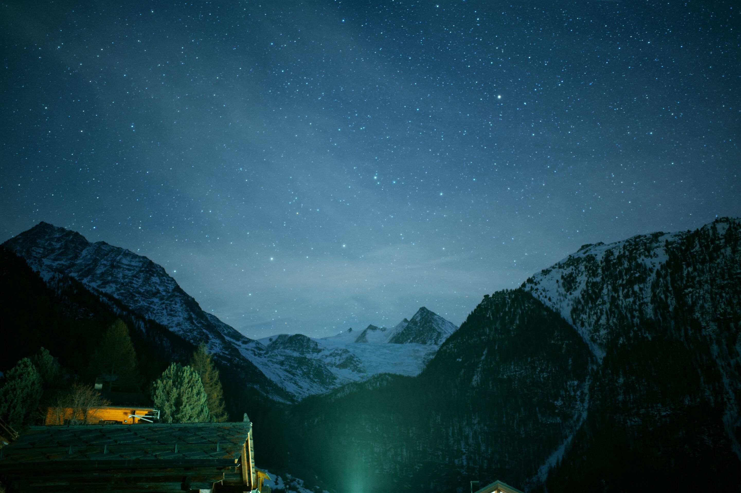 Berglandschap bij nacht met een sterrenhemel, besneeuwde toppen en een gebouw met licht op het dak.