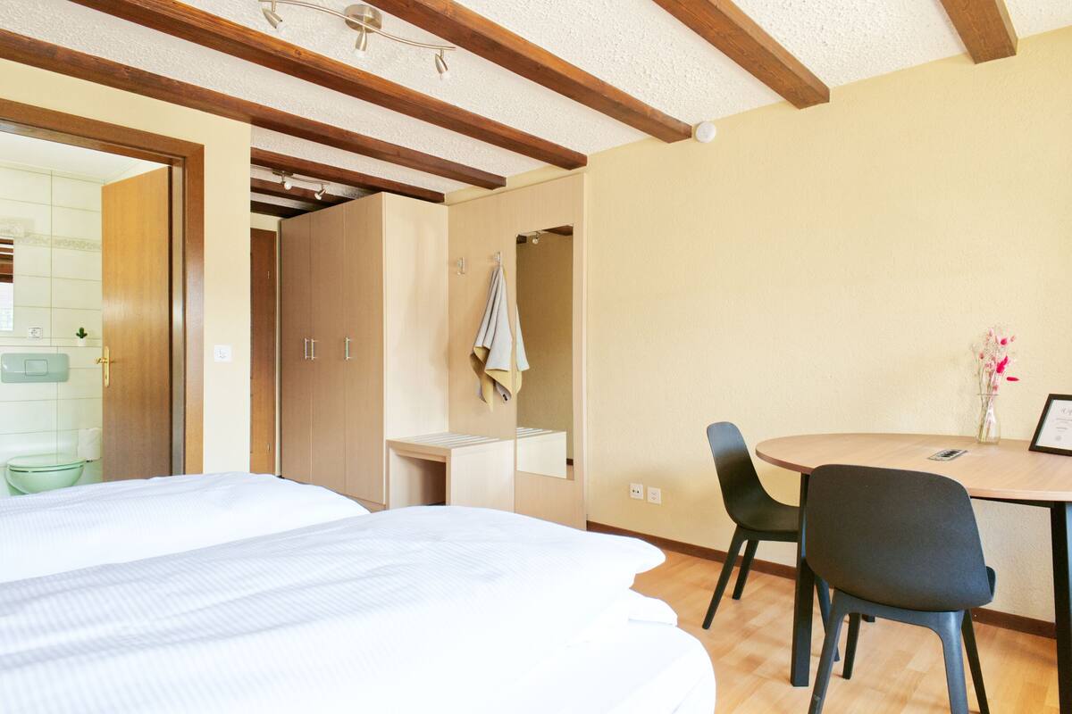 Una habitación de hotel con cama, escritorio, silla y una pequeña mesa con un jarrón.