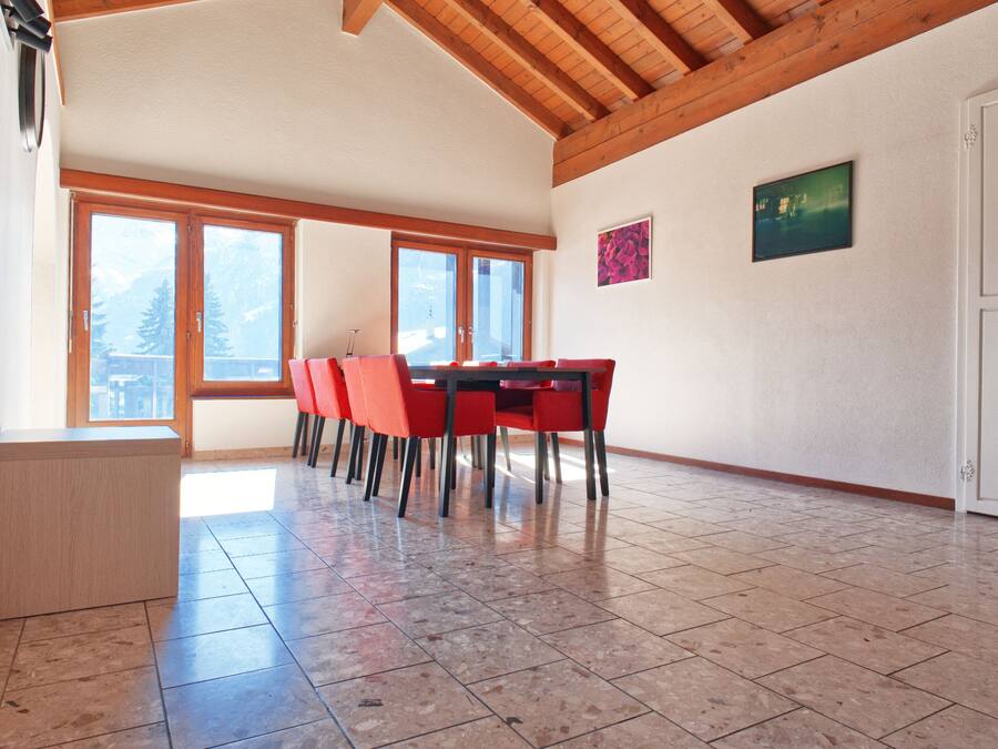 A spacious room with a wooden ceiling, large windows, a dining table with chairs, and a small cabinet.