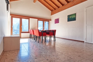 A spacious room with a wooden ceiling, large windows, a dining table with chairs, and a small cabinet. - Alpia FlexiHomes (Graechen)