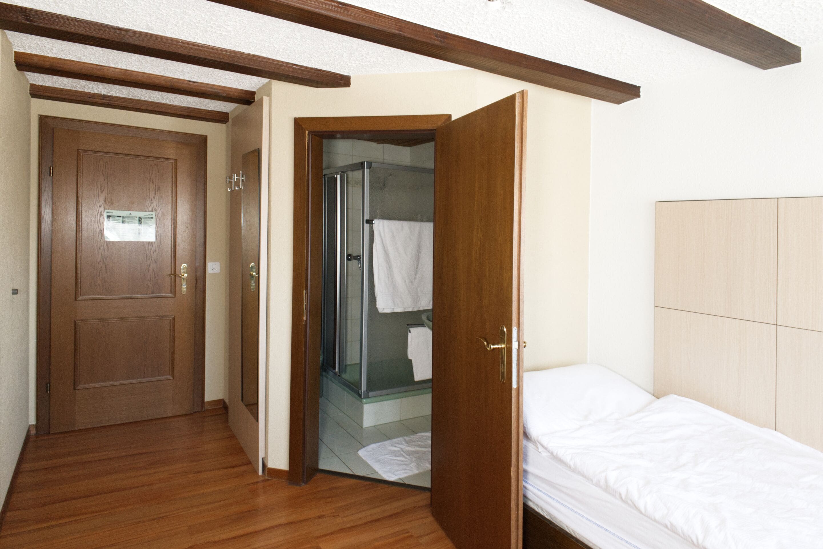 A bedroom with a wooden door, a glass shower, and a bed with white bedding.