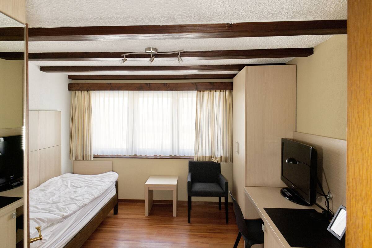 Habitación de hotel compacta con una cama individual, un escritorio pequeño con computadora, una silla y un televisor.
