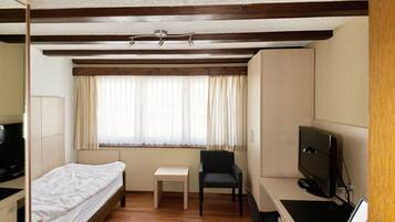 Ein kleines Hotelzimmer mit einem Einzelbett, einem Schreibtisch mit Computer, einem Stuhl und einem Fernseher.