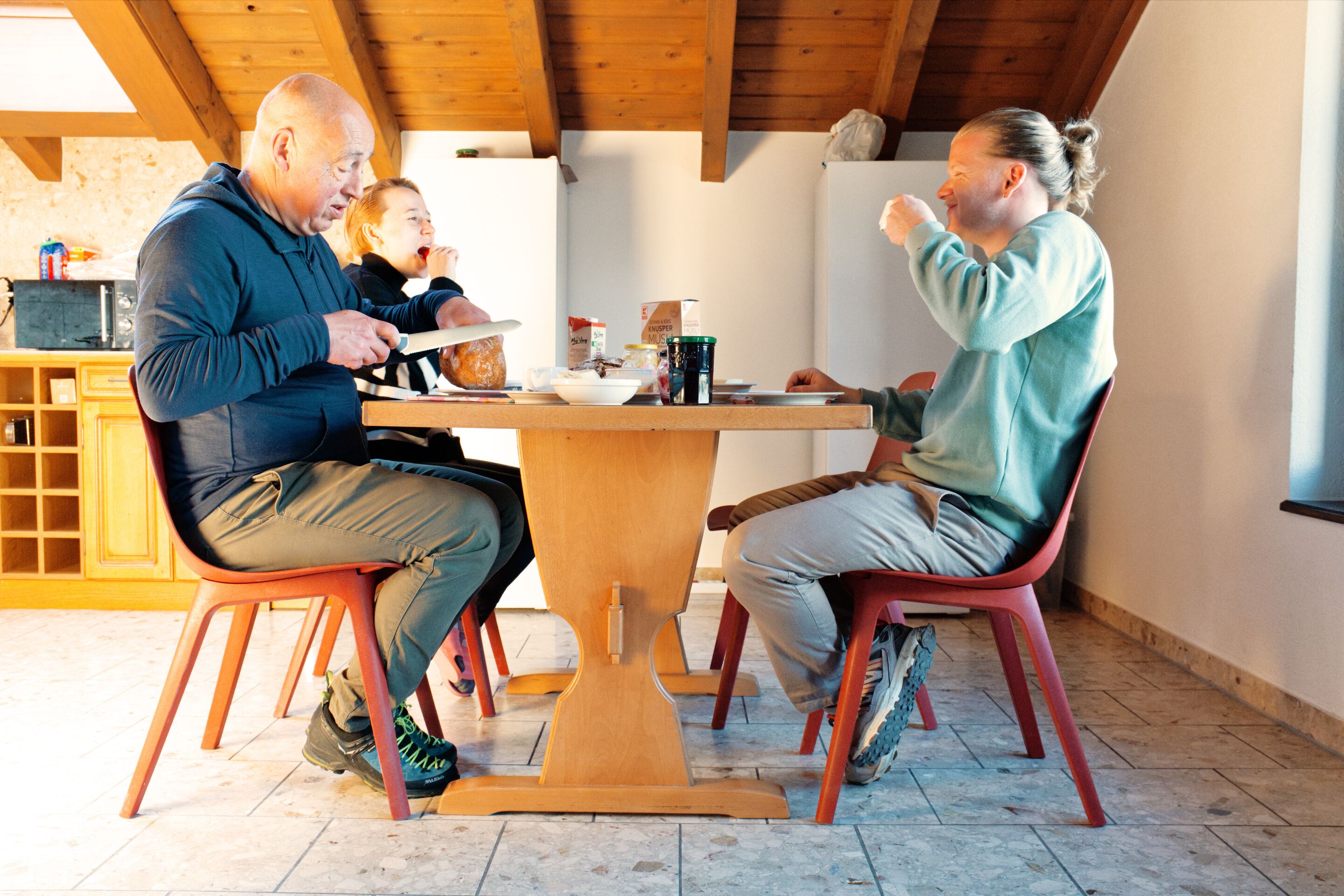 A dining area with a wooden table and chairs, a kitchen in the background, and a window providing natural light.